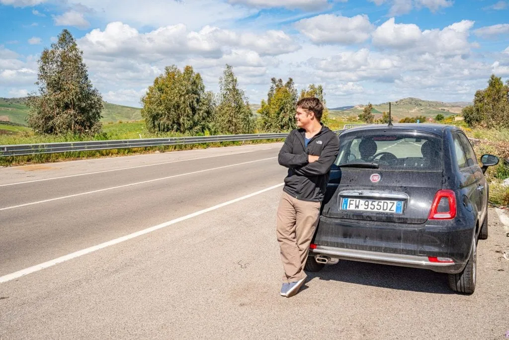 Jeremy Storm leaning against a small black Sicily rental car looking out over an empty road in Sicily