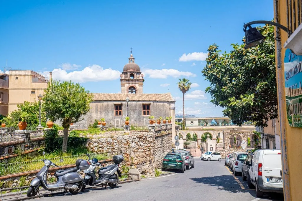 Photo of a street in Taormina Sicily with cars parked on either side. There's a church visible toward the back of the photo.