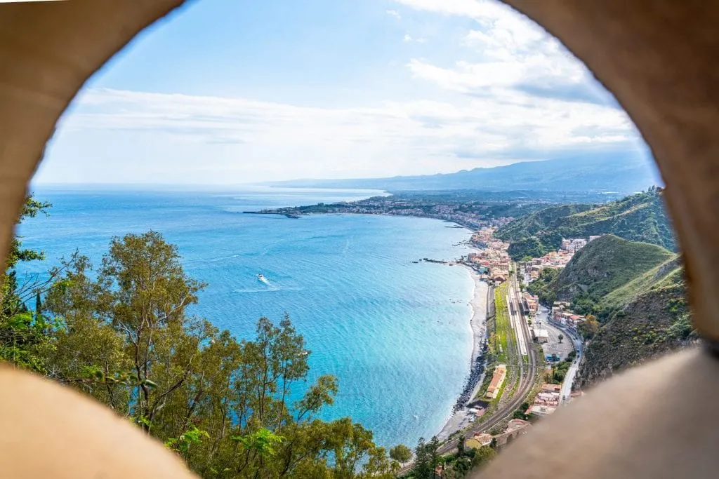 View of a beach along the Ionian Sea as seen by peaking through a gate at Villa Comunale, one of the best places to visit in Taormina Sicily!