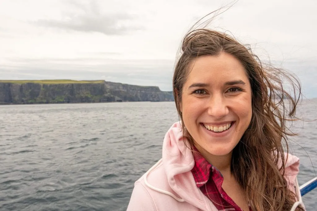 Kate Storm wearing a pink jacket and smiling at the camera during a Cliffs of Moher cruise--the cliffs as visible in the background.