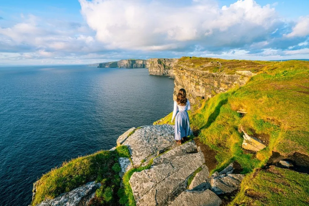 Kate Storm in a blue skirt standing in front of the Cliffs of Moher in Ireland. She's looking away from the camera.
