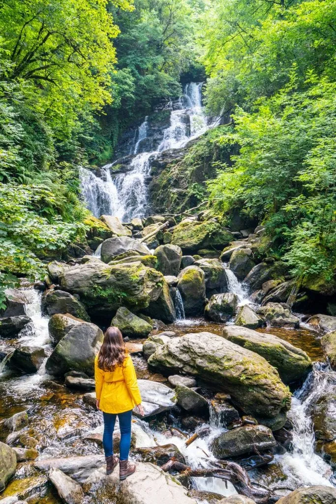 Kate Storm in a yellow raincoat standing in front of Torc Waterfall in Killarney National Park Ireland