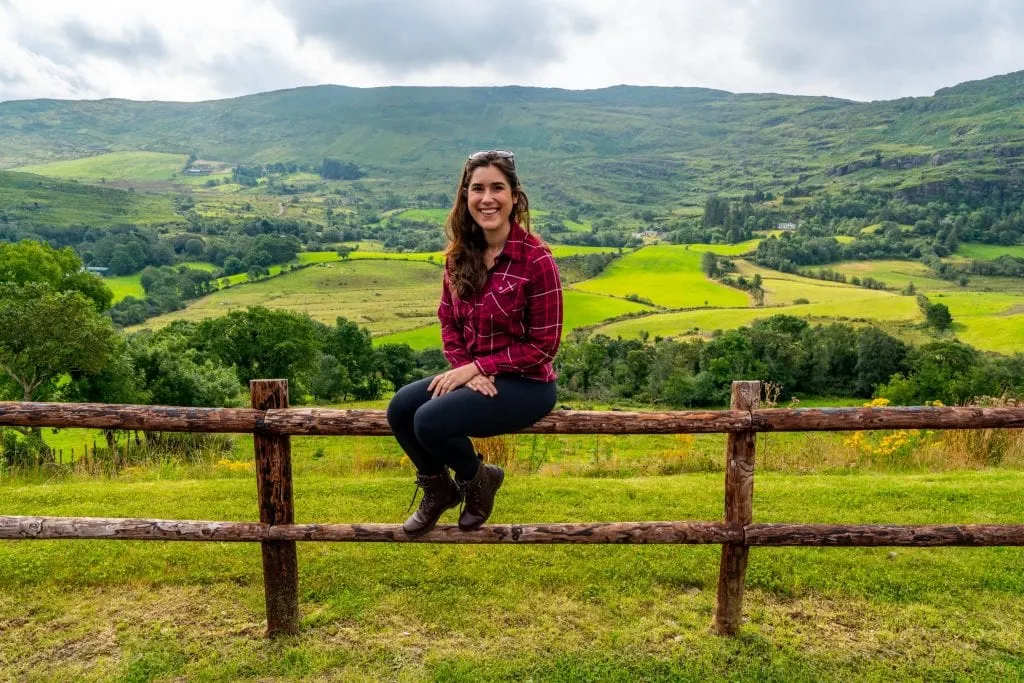 Kate Storm sitting on a wooden split level fence in Ireland with countryside behind her.