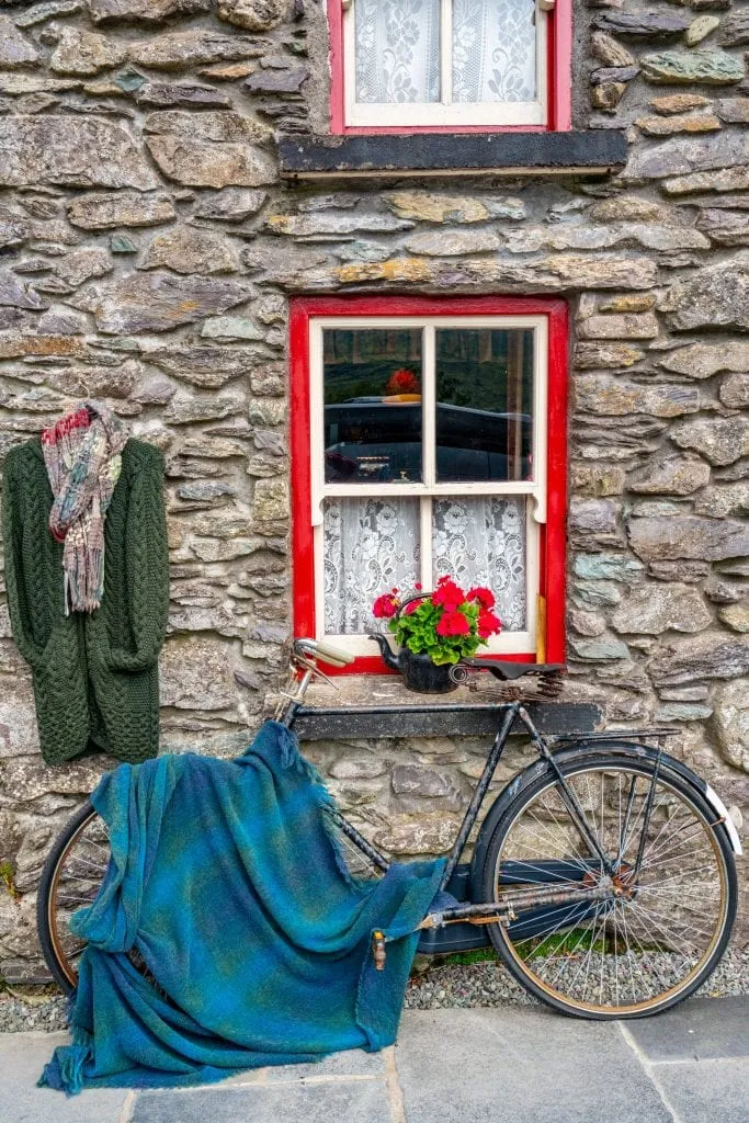 Stone building in Ireland with a bike leaning against the wall. A wool blue blanket is laying over the bike and a window in the building is lined in red