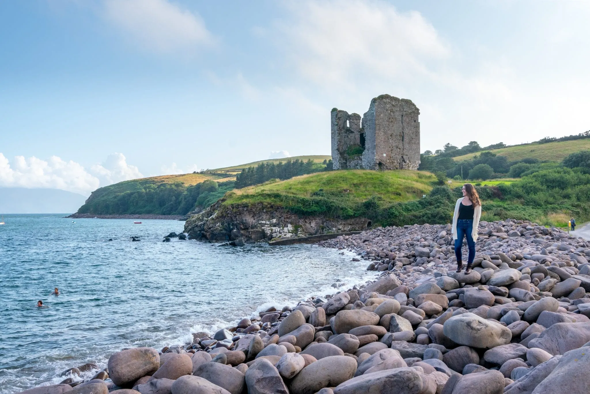 Kate Storm standing on a pebble beach on Dingle Peninsula, Ireland. Minard Castle is behind her. This is a great example of what to wear in Ireland!