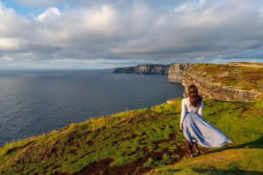 Kate Storm in a blue skirt looking away from the camera and looking out at the Cliffs of Moher--if you plan carefully, views like this won't factor into your trip to Ireland cost, because they're free!