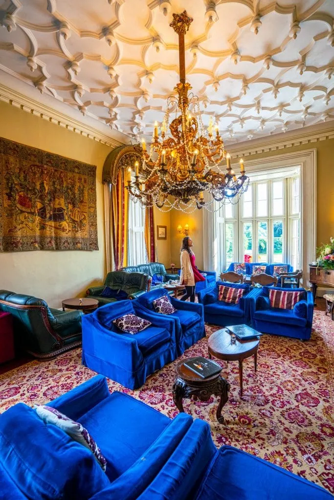 Kate Storm standing in the Library of Belleek Castle County Mayo Ireland, surrounded by blue couches with a chandelier hanging from the ceiling