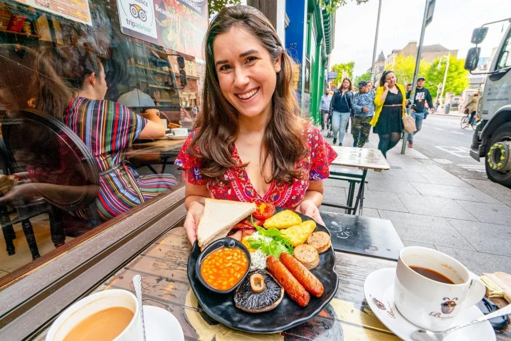 Kate Storm in a red dress holding up a full Irish breakfast while sitting at a small table on the sidewalk in Dublin
