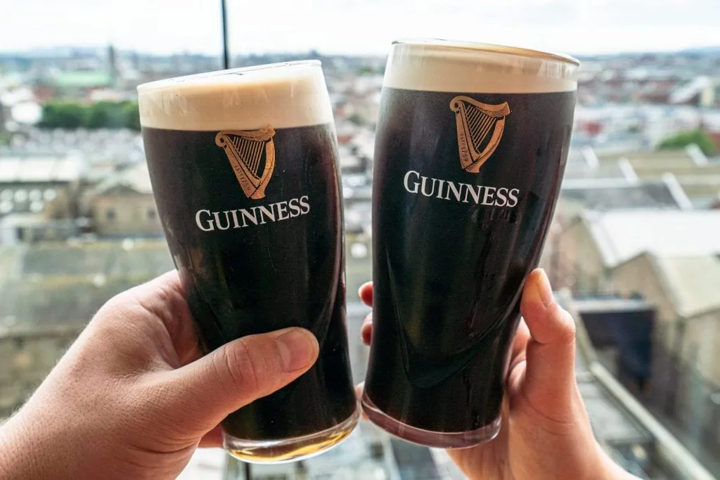 Two pints of Guinness being held up in front of a window at the Guinness Storehouse--definitely pay this spot a visit during your 2 days in Dublin Ireland.