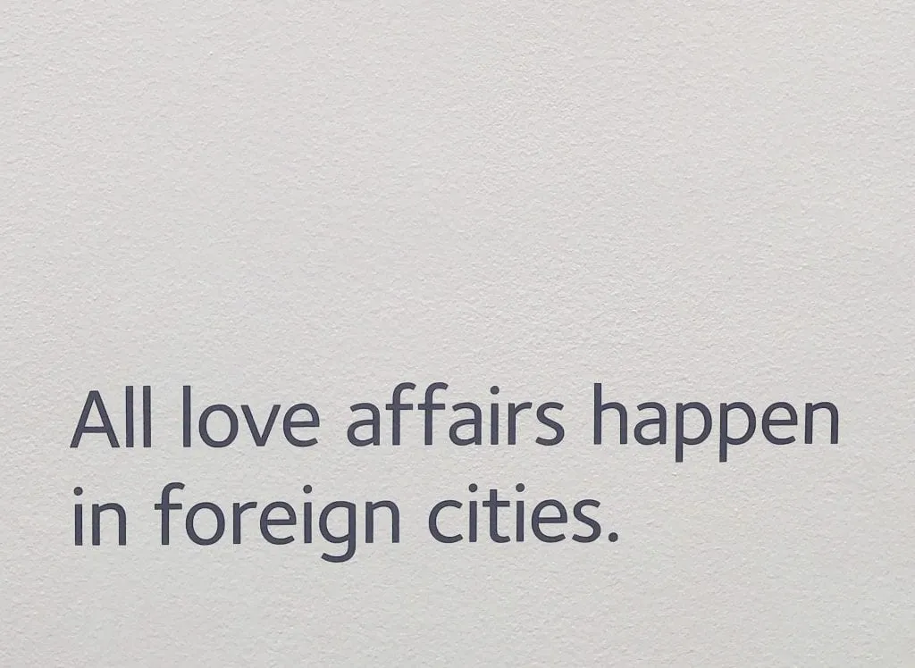 Photo of white wall in the Museum of Broken Relationships in Zagreb Croatia. Black text reads "All love affairs happen in foreign cities."