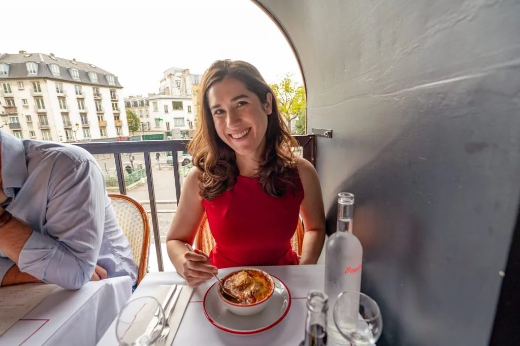 Kate Storm in a red dress with a bowl of French onion soup on the table in front of her--Bouillon Pigalle is a great pick for lunch during your weekend trip to Paris!