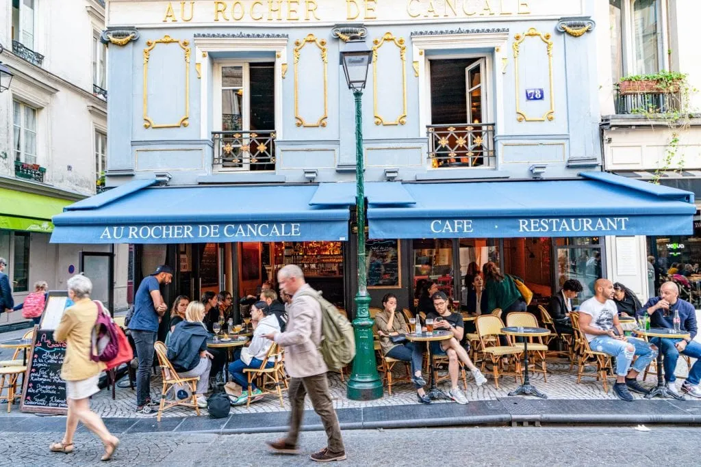 Blue building with a blue awning of a cafe in Paris with a man walking in front of it--scenes like this have inspired many quotes about Paris!