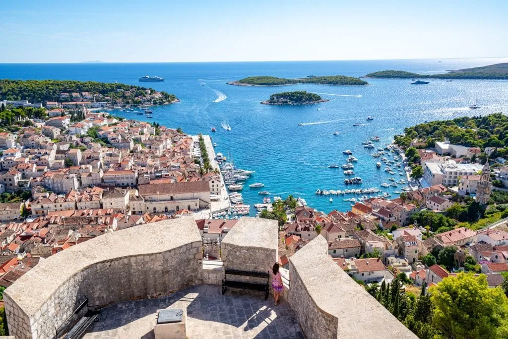 View of Hvar Town from Spanish Fortress with Pakleni islands in background and Kate Storm in a pink dress standing on the fort. Don't miss this view during your 10 day Croatia itinerary!