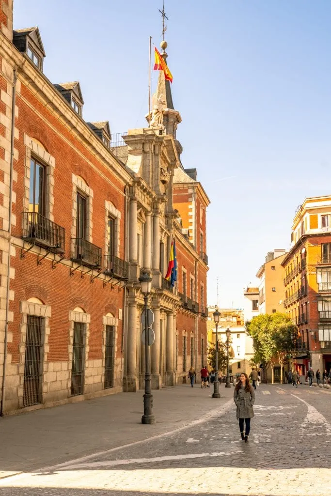 Kate Storm in a gray coat walking down the street in Madrid with a red brick building to her left