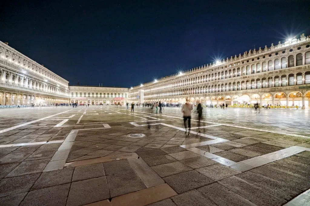 Photo of Piazza San Marco at night in Venice with a few people walking around and lights on the buildings illuminating the square