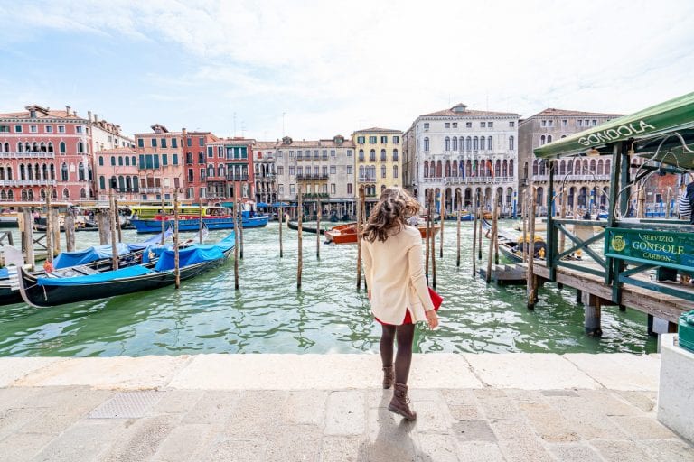 Kate Storm standing with her back to the camera along the Grand Canal, a must-see during a Florence to Venice day trip! Gondolas are parked along the canal and Kate is wearing a cream sweater.