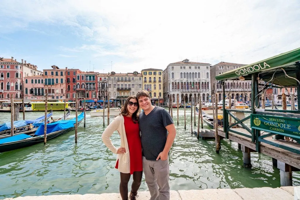 Kate Storm and Jeremy Storm standing along the Grand Canal of Venice. Kate is in a red dress and there are gondolas behind them.