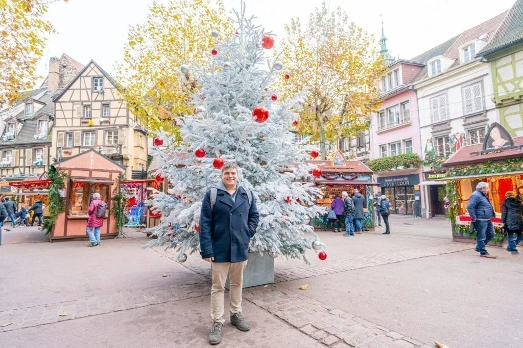 Jeremy Storm in a navy peacoat standing in front of a white Christmas tree during winter in Colmar France