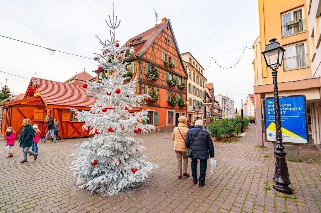 Older couple walking past a white Christmas tree in Colmar with half-timbered houses in the background