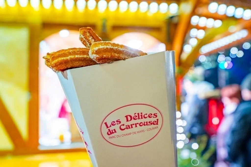 Churros in a white box being held up at a lit up Christmas market stand in Colmar in Winter