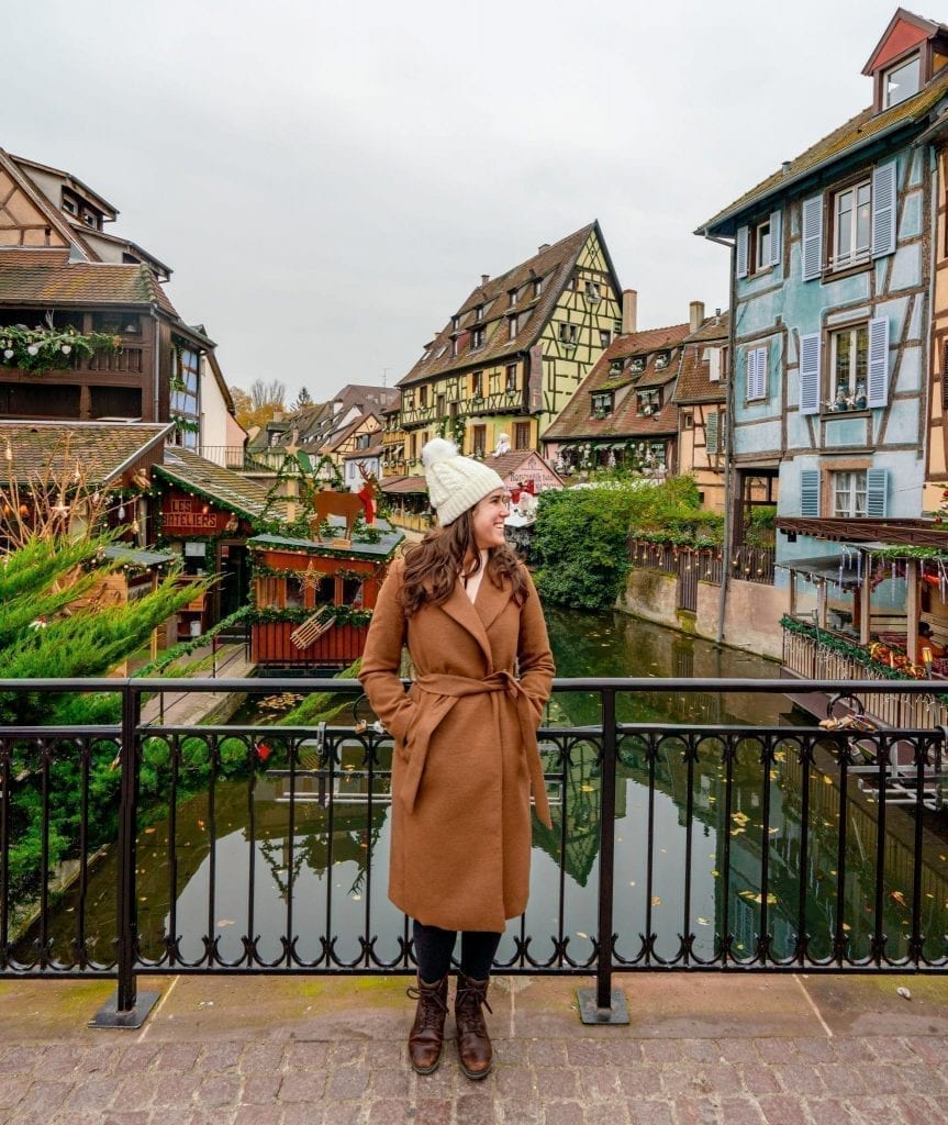 Kate Storm in a brown coat standing in front of a canal in Colmar in winter