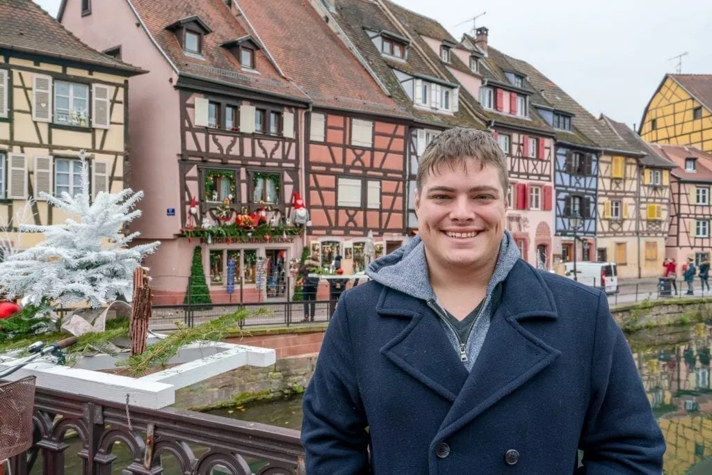 Jeremy Storm in a blue coat standing in front of colorful houses in Colmar France