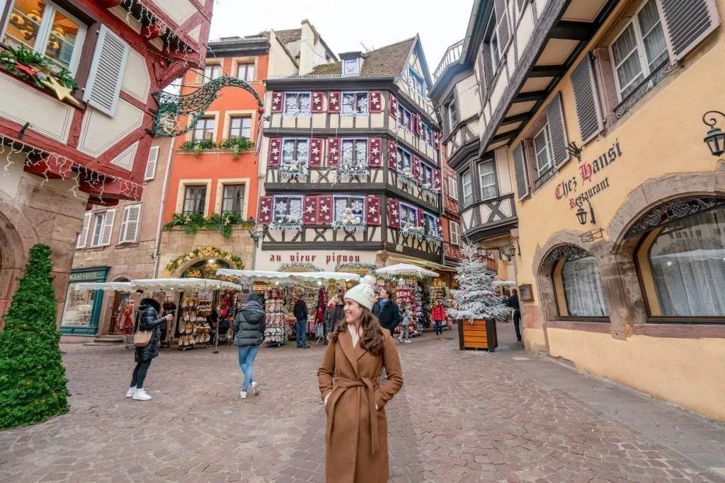 Kate Storm in a brown coat approaching the camera. Several houses decorated for Christmas in Colmar France are behind her.