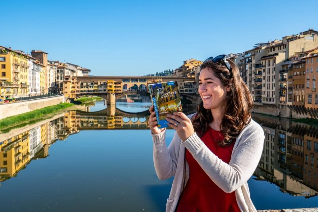 Photo of Kate Storm reading a copy of Moon Florence & Beyond with the River Arno and Ponte Vecchio in the background