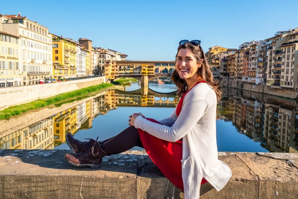 Kate Storm in a red dress sitting on a bridge in front of the Arno with the Ponte Vecchio visible in the background--be sure to seek out this view during your 2 days in Florence Italy