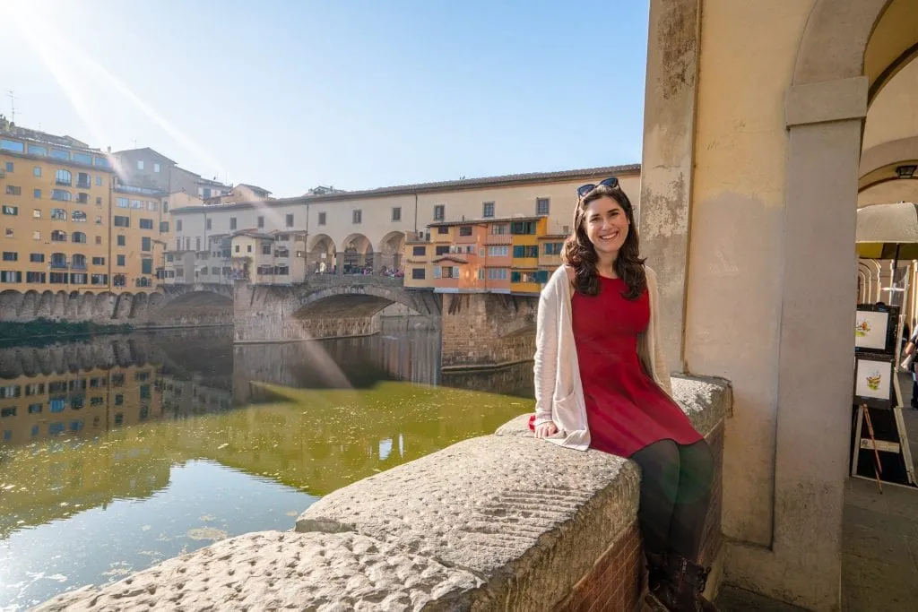 Kate Storm in a red dress sitting on the ledge of a wall with the Ponte Vecchio in the background--these are the kinds of views waiting for you once you finish your drive from Rome to Florence