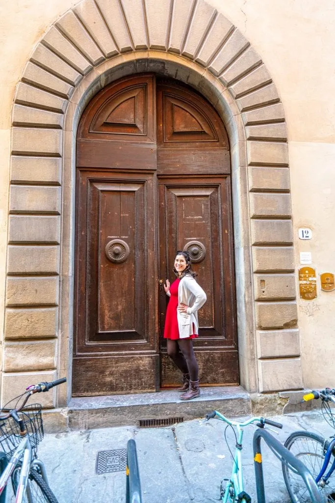 Kate Storm in a red dress entering a large wooden door in Florence