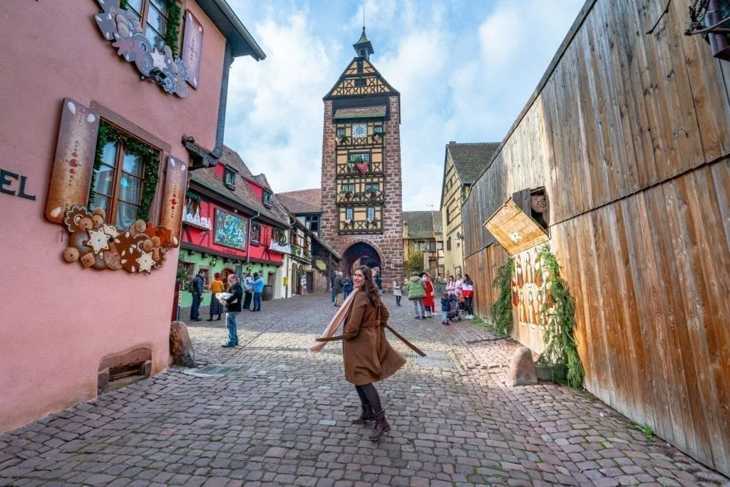 Kate Storm in a brown coat in the Alsace village of Riquewihr with a clock tower in the background