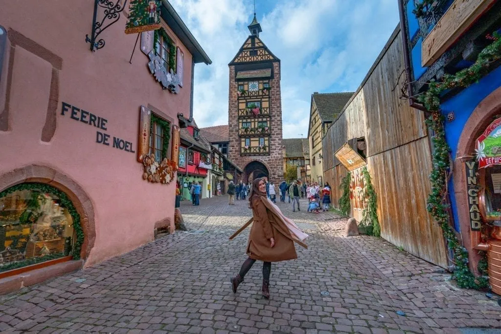 Kate Storm in a brown coat in the Alsace village of Riquewihr with a clock tower in the background