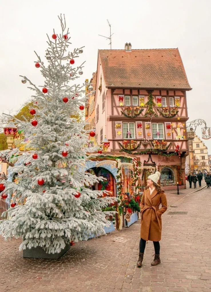 Kate Storm in a brown coat standing with a white Christmas tree in Colmar. A decorated pink house is in the background--this is a classic photo spot in Colmar in winter!