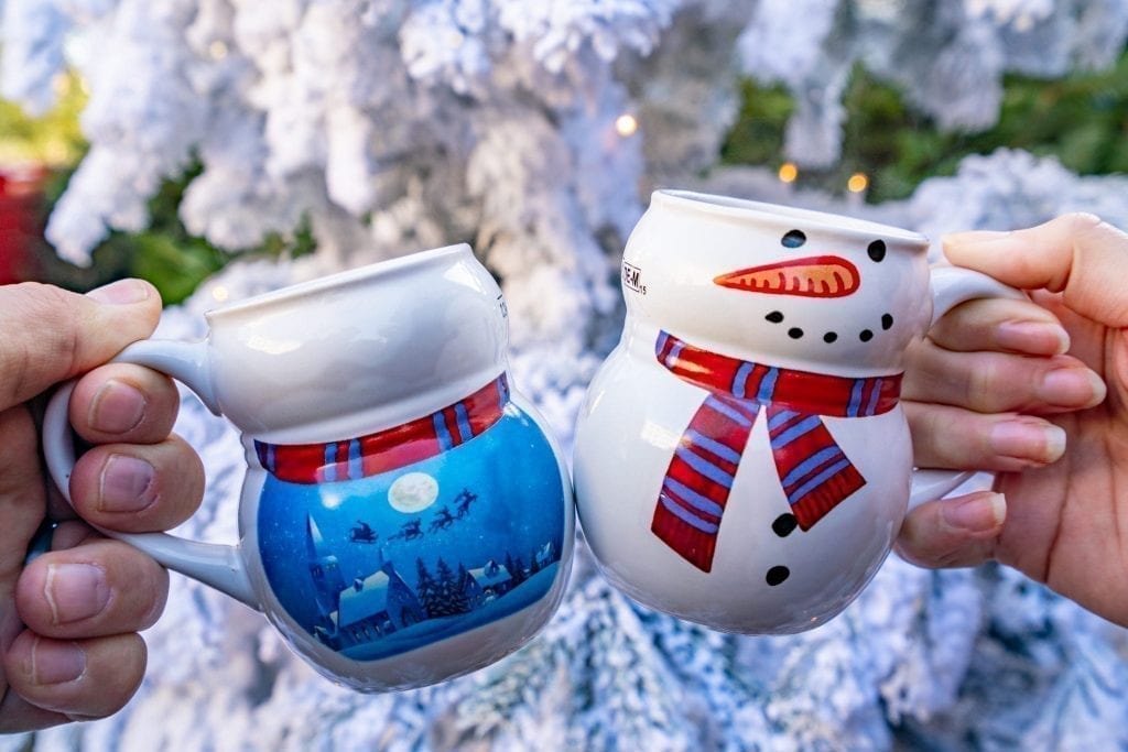 two santa mugs being held up in front of a white christmas tree, one of the best souvenirs christmas markets