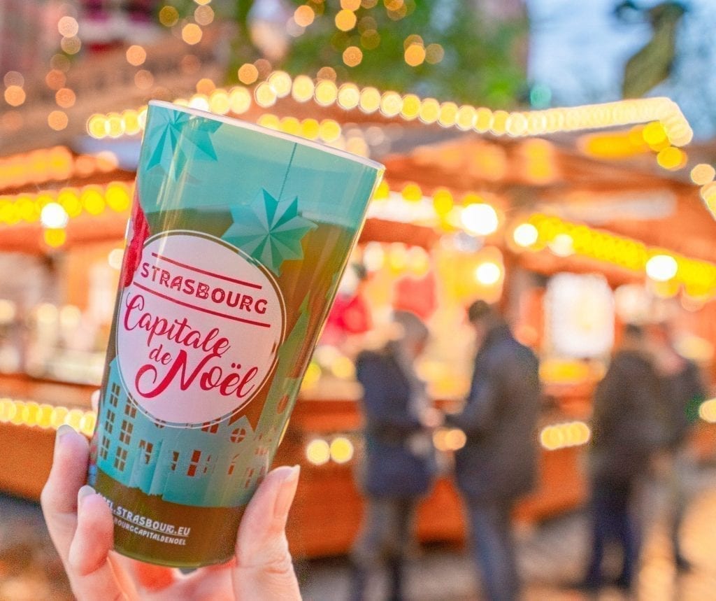 Green cup with red writing stating "Strasbourg Capital de Noel" being held up in front of a Strasbourg Christmas market stall