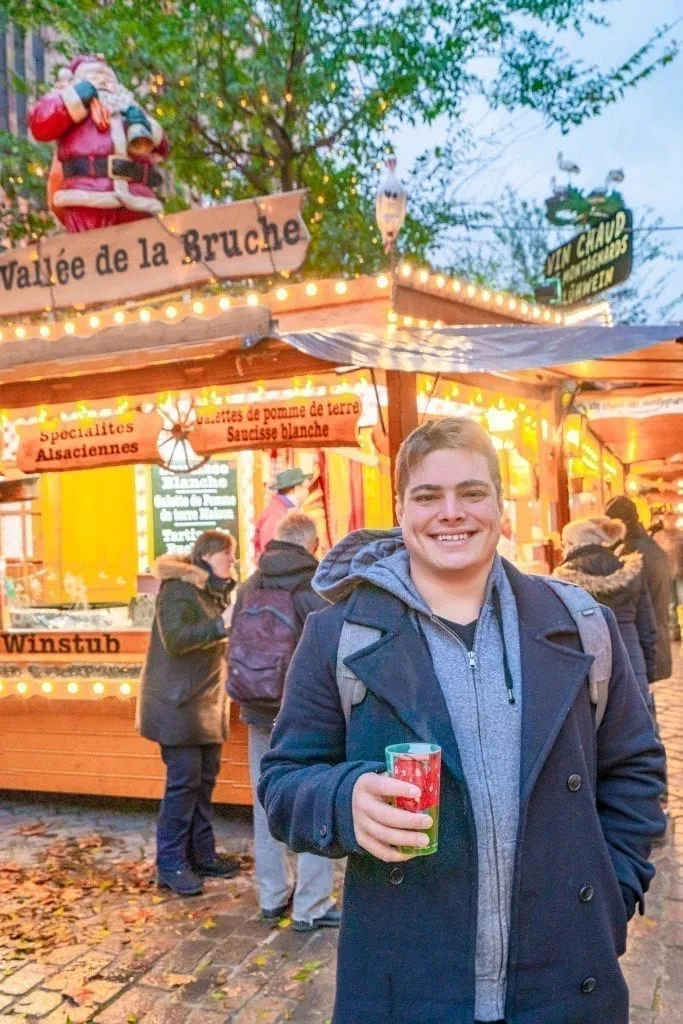 Jeremy Storm in a blue coat holding gluhwein and standing in front of a Strasbourg Christmas market stall