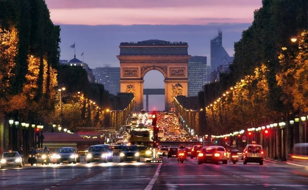 Champs Elysses during the evening in Paris, with traffic in front, Arc de Triomphe in the rear of the photos, and a slight pink sunset visible.