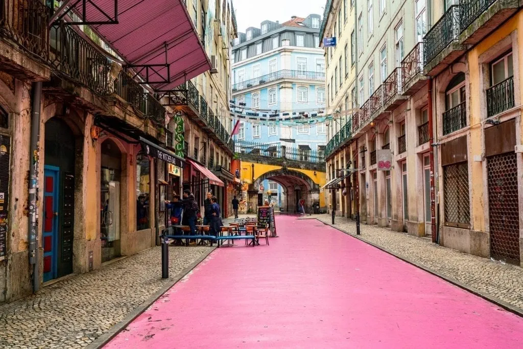 Lisbon Pink Street empty during the day with yellow bridge and blue building visible in the background. One of the best things to see in Lisbon Portugal