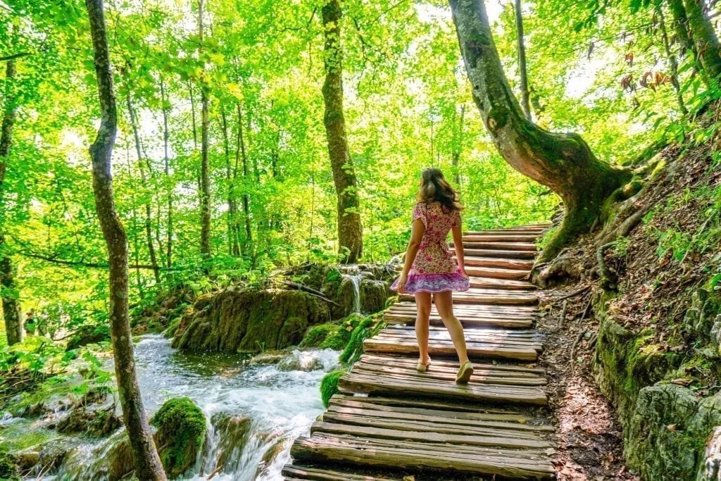 Kate Storm in a pink dress twirling on a boardwalk when visiting Plitvice Lakes Croatia. Trees surround the boardwalk and water is running under it.