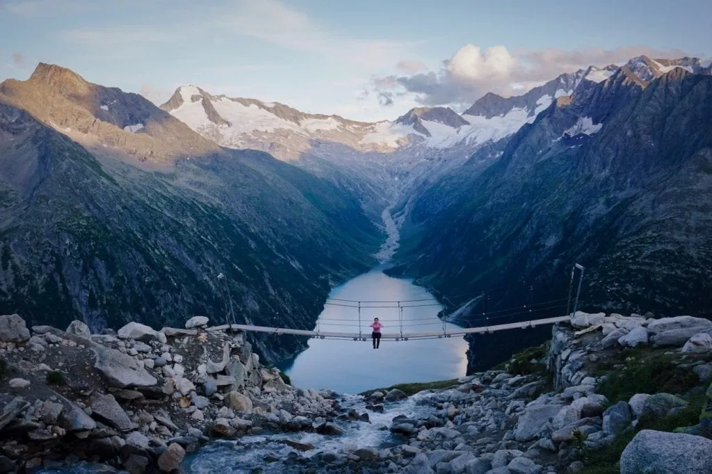 Person sitting on a small footbridge in Zillertal Alps in Austria, with a lake and mountain range visible in the distance