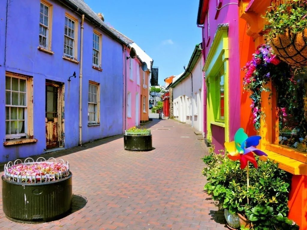 Brightly colored street in Kinsale Ireland with a purple building in the left foreground and a red building on the right foreground. Kinsale is one of the prettiest small towns in Ireland.