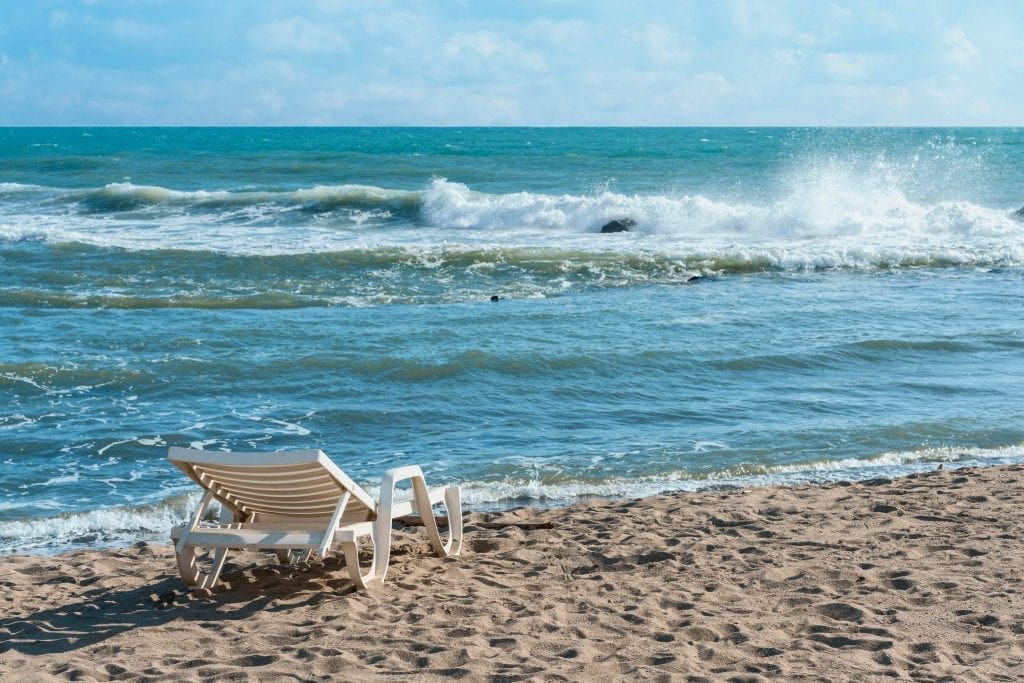 White deck chair on the edge of Ostia Lido beach with the sea and one crashing wave taking up most of the photo