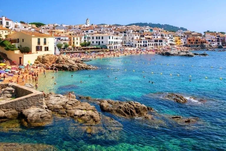 coastline of Costa Brava Spain as seen from across with water, with a village visible in the distance. Spain's Costa Brava is one of the best European road trip routes!