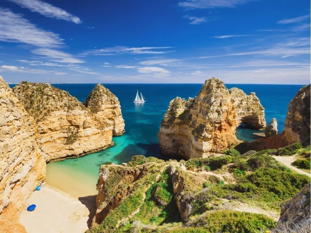 View of small beach on Algarve Coast in Portugal with a sailboat in the distance and rocky cliffs jutting out to sea