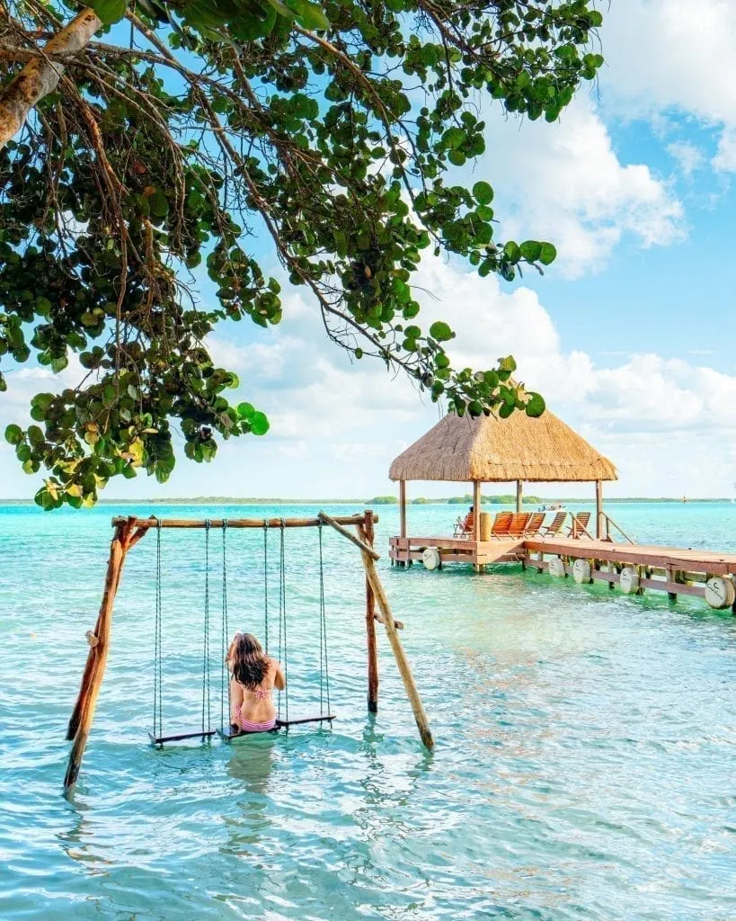 Kate Storm sitting on a swing in the water in Bacalar Mexico wearing a pink bikini--plenty of bathing suits definitely belong on your beach vacation packing list