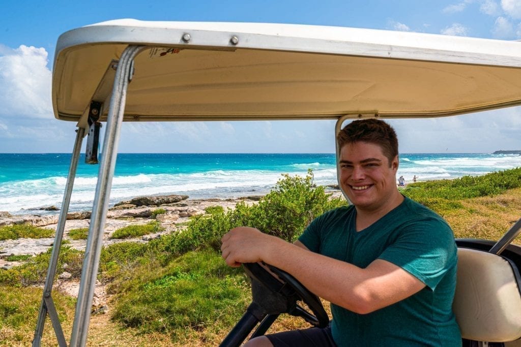 Jeremy Storm in a green shirt sitting behind the wheel of a golf cart with the east coast of Isla Mujeres in the background. Renting a golf cart is one of the best things to do in Isla Mujeres