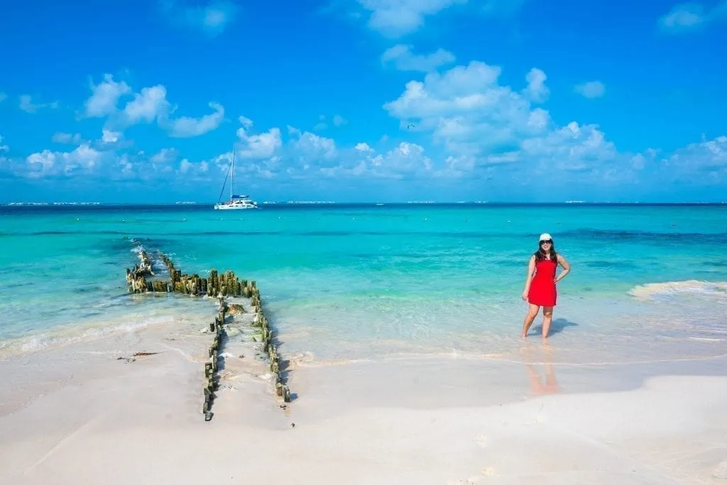 Kate Storm in a red dress standing on Playa Norte in Isla Mujeres. The remains of a dock are to her left.
