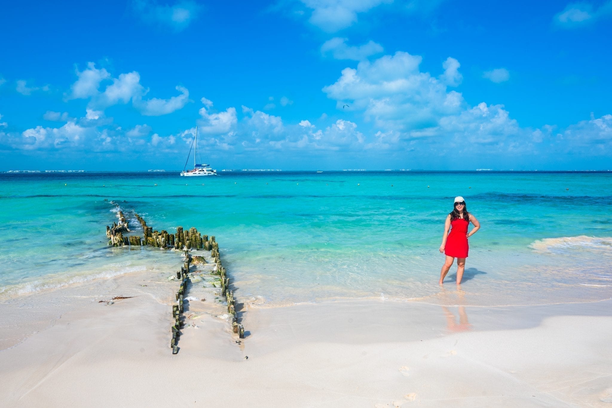 Kate Storm in a red dress standing on Playa Norte in Isla Mujeres. The remains of a dock are to her left.