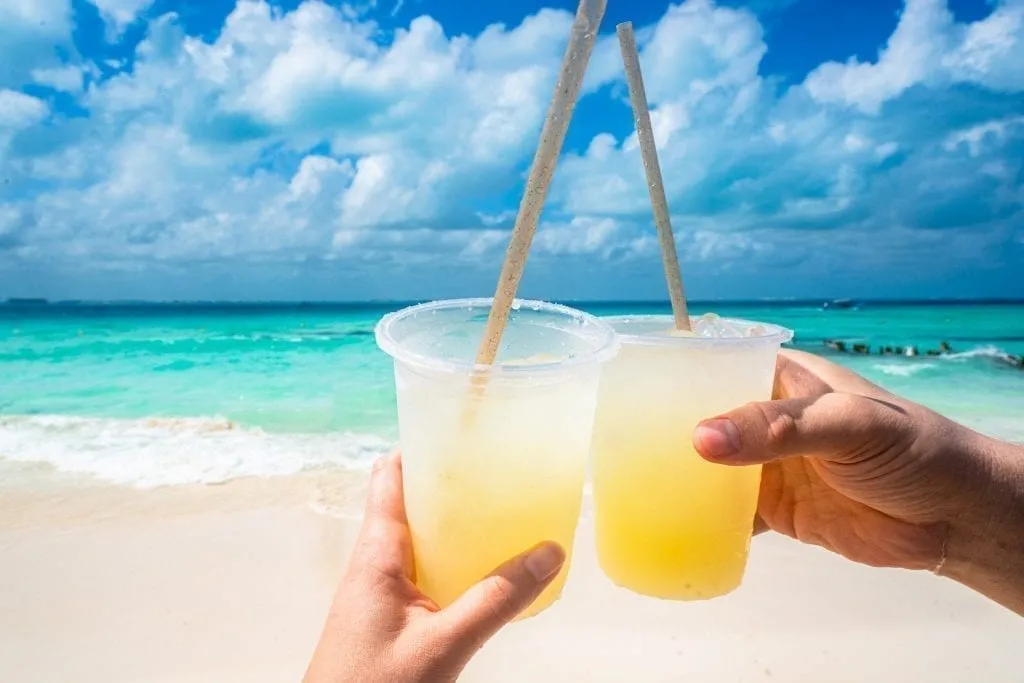 two margaritas being held out in front of the beach--relaxing here belongs on any list of what to do in isla mujeres mexico